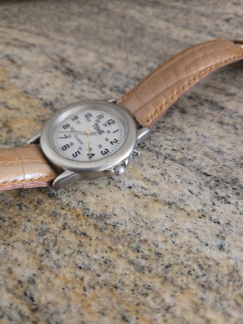 May include: A silver-toned wristwatch with a white face and black numerals. The watch has a light brown leather band. The watch face reads "TIME QUARTZ".