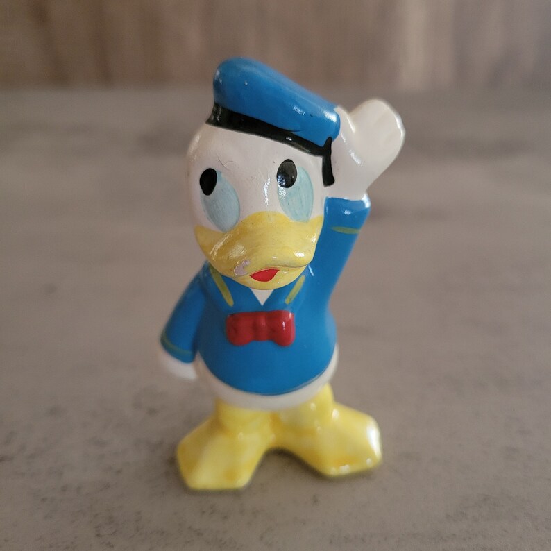 Op de afbeelding: Een keramiek beeldje van Donald Duck met een blauwe matrozenpet en een blauw-wit gestreept matrozenoverhemd. Hij heeft een gele snavel en witte ogen.