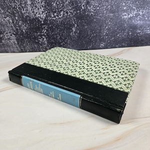 Op de afbeelding: Een hardcover boek met een lichtgroene en witte patrooncover en een zwarte rug. De titel van het boek is zichtbaar op een blauw ruglabel. Het boek ligt op een wit en grijs oppervlak.