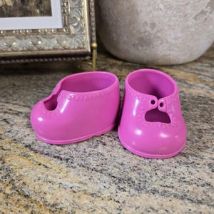 Puede incluir: Un par de zapatos de muñeca de plástico rosa brillante con punta redondeada, abertura baja y un patrón triangular moldeado a lo largo del cuello. Los zapatos tienen un acabado liso y brillante y un detalle recortado distintivo en la parte delantera.