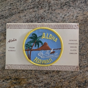 Può includere: Un'applicazione rotonda, blu e gialla di Aloha Hawaii. L'applicazione presenta una palma, una montagna e un surfista. Il testo "Aloha Hawaii" è ricamato in giallo. L'applicazione è su una carta decorativa.