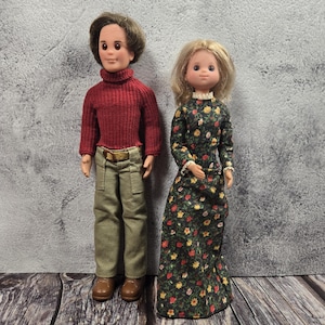 May include: Two vintage dolls, a man and a woman, stand side-by-side. The man doll wears a red turtleneck sweater, khaki pants, and brown shoes. The woman doll is in a floral print dress with a white lace collar. The dolls are on a wooden surface.