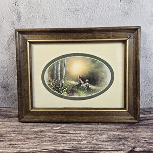 May include: A framed oval print featuring a forest scene with two figures. The print is set within a gold-toned wooden frame. The artwork depicts a sunlit clearing with trees and figures, creating a serene, pastoral aesthetic.