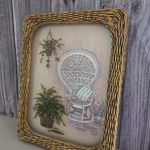 May include: A wicker framed picture with a print of a white wicker chair with a green and white striped pillow. The chair is surrounded by potted plants.