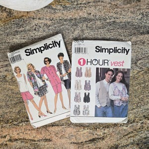 May include: Two vintage Simplicity sewing pattern envelopes. One features illustrations of women in various outfits, and the other showcases a vest pattern with the text "1 HOUR vest". Both are on a textured surface.
