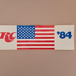 May include: A white sticker with a red, white, and blue American flag in the center. The sticker also has the text "RC" in red and white on the left side and "'84" in blue on the right side.