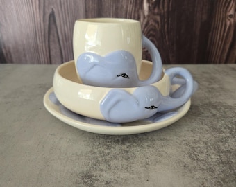 May include: A cream-colored ceramic teacup, saucer, and bowl set. The set features a light blue elephant design on each piece. The elephant's trunk forms the handle of the cup and the bowl's rim. The set is on a gray surface.