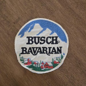 Puede incluir: Un parche redondo blanco bordado con una escena de montaña azul y el texto "Busch Bavarian".
