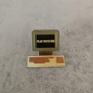Peut inclure: Épingle en forme d'ordinateur avec l'inscription "PLAY WITH ME" sur l'écran. L'épingle a un clavier marron et un écran bordé de vert. Le texte est en lettres dorées sur fond noir. L'épingle est en métal.