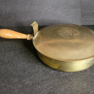 Pode incluir: Um cinzeiro de latão com uma pega de madeira. O cinzeiro tem um design decorativo na parte superior.