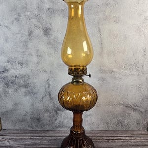 Może przedstawiać: Bursztynowa szklana lampa naftowa z dekoracyjną podstawą i kloszem. Lampa posiada wysoki, rozszerzany komin i okrągły, wzorzysty zbiornik. Podstawa jest w ciemniejszym odcieniu brązu. Lampa ma estetykę vintage.