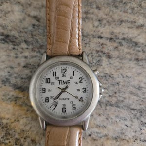 May include: A silver-toned quartz watch with a white face and a light brown leather band. The watch face displays the word "TIME" and the numbers 1 through 24. The band has a textured, crocodile-like pattern.