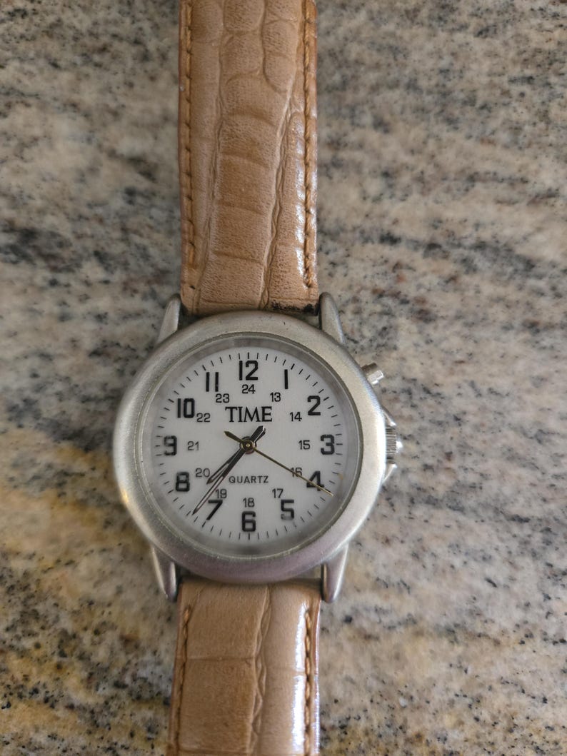 May include: A silver-toned quartz watch with a white face and a light brown leather band. The watch face displays the word "TIME" and the numbers 1 through 24. The band has a textured, crocodile-like pattern.