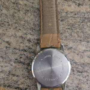 May include: A stainless steel quartz watch with a light brown leather strap. The watch back is visible, with text indicating "Japan Movt" and "Hong Kong". The watch has a silver-colored case.