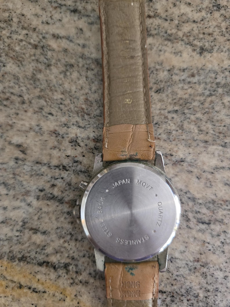 May include: A stainless steel quartz watch with a light brown leather strap. The watch back is visible, with text indicating "Japan Movt" and "Hong Kong". The watch has a silver-colored case.