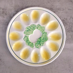 May include: A white ceramic egg plate with 12 yellow egg-shaped indentations. The plate has a green leafy design in the centre.