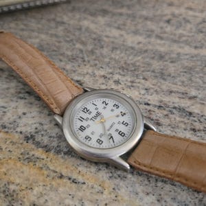 May include: A silver-toned quartz watch with a white face and black numerals. The watch has a light brown leather band with a crocodile pattern. The watch face reads "TIME QUARTZ".