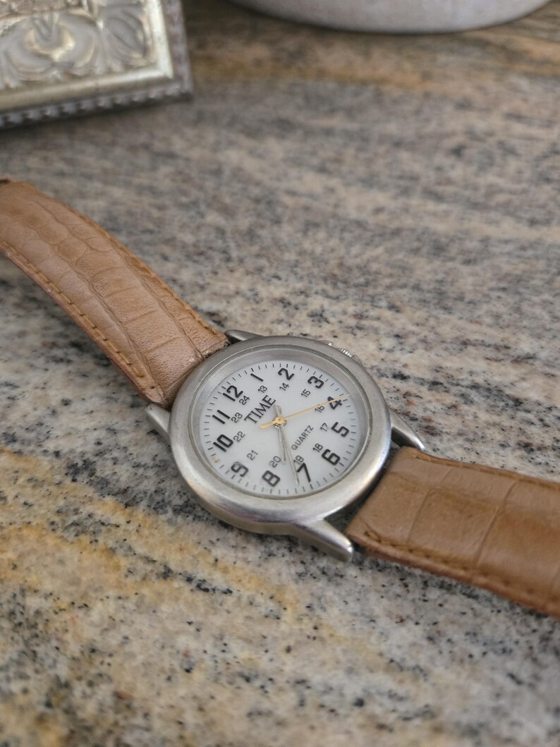May include: A silver-toned quartz watch with a white face and black numerals. The watch has a light brown leather band with a crocodile pattern. The watch face reads "TIME QUARTZ".