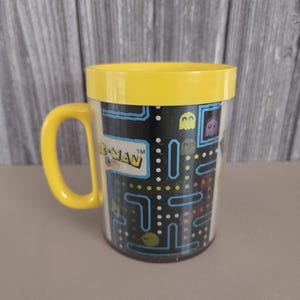 May include: Yellow Pac-Man mug with a black and white Pac-Man maze design. The mug has a yellow handle and the text "Pac-Man" is printed on the side.