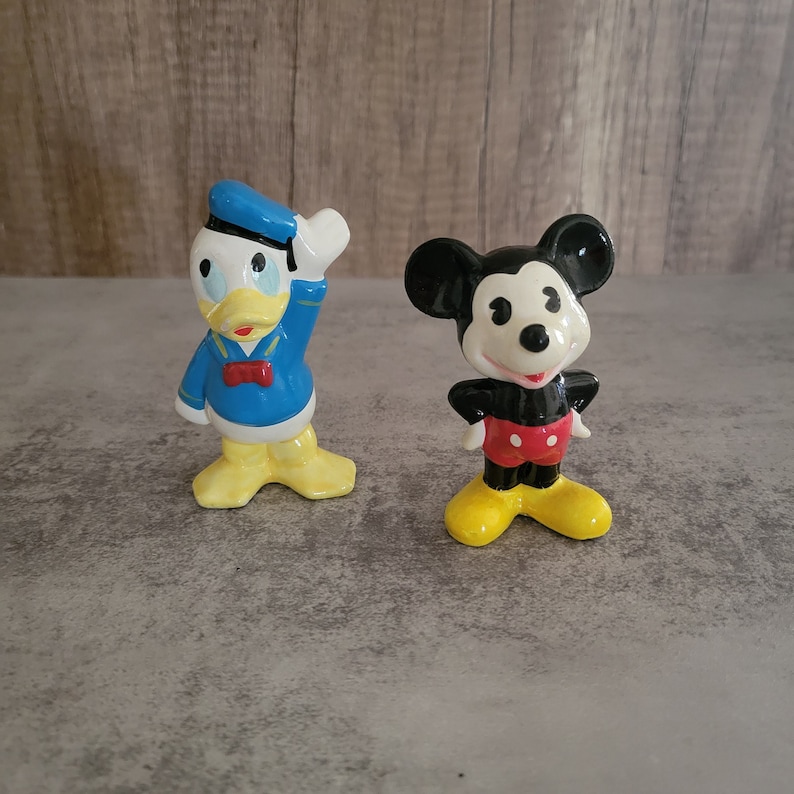 Op de afbeelding: Een vintage set van keramische beeldjes met Mickey Mouse en Donald Duck. Mickey Mouse draagt zijn kenmerkende rode broek en gele schoenen, terwijl Donald Duck een blauw matrozenoverhemd en gele schoenen draagt.