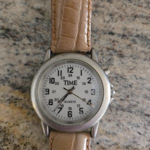 May include: A silver-toned quartz watch with a white face and black numerals. The watch has a light brown leather band. The watch face displays the word "TIME".
