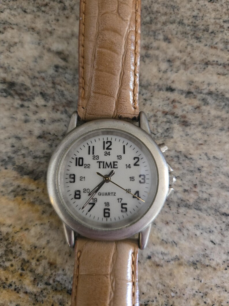 May include: A silver-toned quartz watch with a white face and black numerals. The watch has a light brown leather band. The watch face displays the word "TIME".