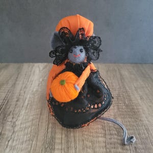 May include: A small gray felt mouse wearing a black lace dress and an orange pumpkin hat, holding an orange felt pumpkin.