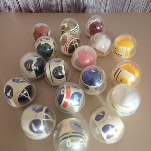 Peut inclure: Une collection de 18 casques de football miniatures dans des étuis en plastique transparent. Les casques sont décorés avec des logos et des couleurs d'équipe, notamment le rouge, le blanc, le bleu, le vert, le jaune et le noir.