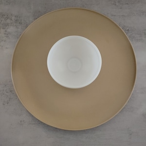 May include: A white ceramic bowl sits in the center of a large, round, beige serving tray. The tray and bowl are both empty.