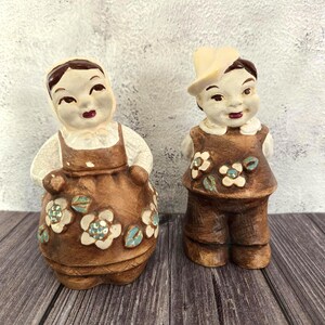 May include: A pair of vintage ceramic figurines depicting a boy and a girl. The figures are dressed in brown outfits with floral accents. The girl wears a dress and the boy wears a hat. The figurines have painted facial features.