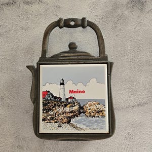 May include: A metal trivet in the shape of a teapot, featuring a ceramic tile with a coastal scene of a lighthouse and the word "Maine". The trivet has a handle and a decorative design.