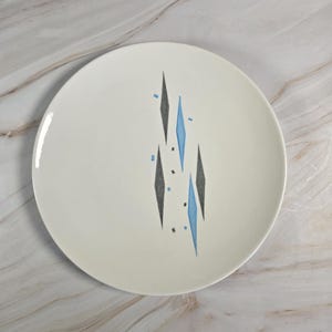 Peut inclure: Une assiette en céramique blanche avec un design moderne du milieu du siècle. L'assiette présente un motif de losanges noirs et bleus. L'assiette est ronde et a une finition brillante. L'assiette est adaptée pour servir des aliments.