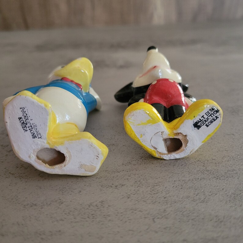 Op de afbeelding: Onderkant van twee keramische beeldjes, een van Mickey Mouse en een van Donald Duck. Beide beeldjes zijn geel, wit, rood en blauw. Het Mickey Mouse beeldje heeft de tekst "Walt Disney Productions Korea" op de onderkant. Het Donald Duck beeldje heeft de tekst "Walt Disney Productions Korea" op de onderkant.