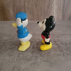 Op de afbeelding: Een vintage set keramische beeldjes met Mickey Mouse en Donald Duck. Mickey draagt een rode broek en gele schoenen, terwijl Donald een blauw matrozenpak en gele schoenen draagt.