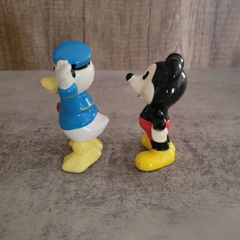 Op de afbeelding: Een vintage set keramische beeldjes met Mickey Mouse en Donald Duck. Mickey draagt een rode broek en gele schoenen, terwijl Donald een blauw matrozenpak en gele schoenen draagt.