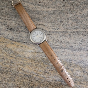 May include: A silver-toned quartz watch with a white face and black numerals. The watch has a brown, textured leather band. The watch face reads "TIME" and "QUARTZ".