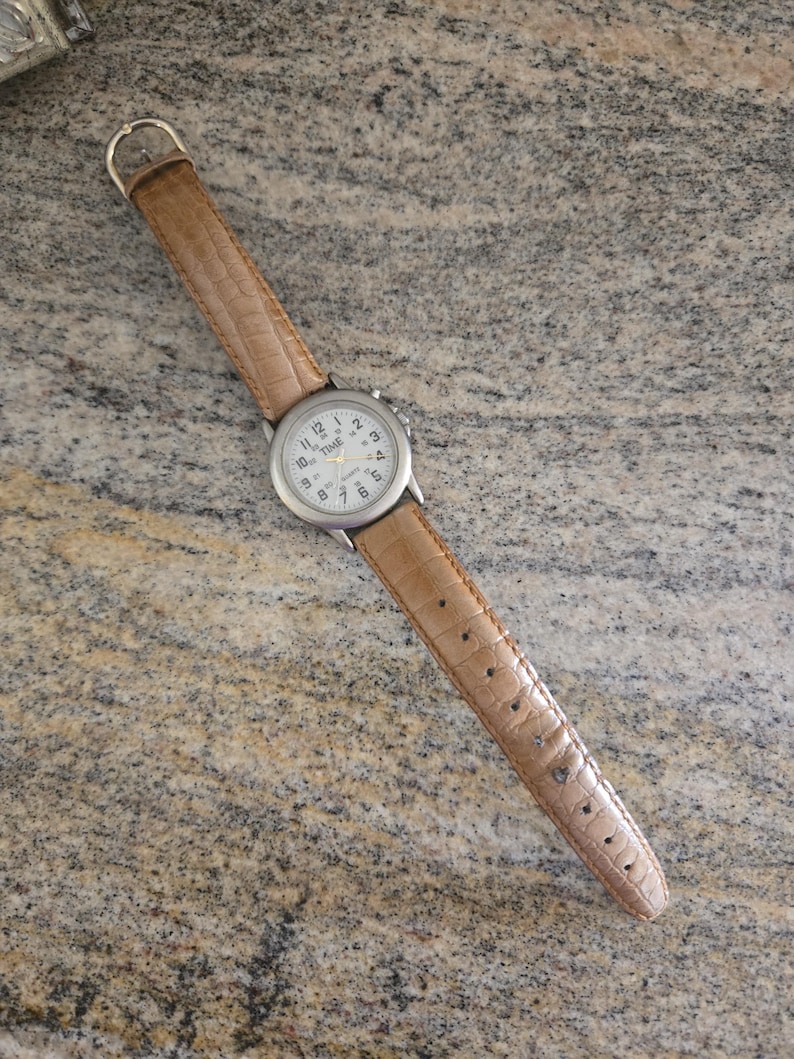 May include: A silver-toned quartz watch with a white face and black numerals. The watch has a brown, textured leather band. The watch face reads "TIME" and "QUARTZ".
