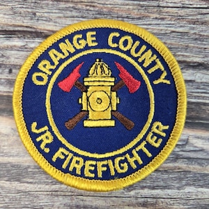 May include: A circular patch with a blue background and gold trim. The patch reads "ORANGE COUNTY JR. FIREFIGHTER" in gold lettering. The center features a gold fire hydrant with crossed brown axes and red axe heads.
