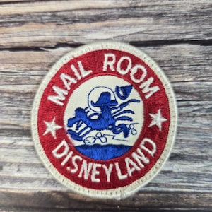 May include: Round patch with a red border and white center. The text "MAIL ROOM DISNEYLAND" is in red. A blue cartoon character is in the center. Two white stars are on the border.
