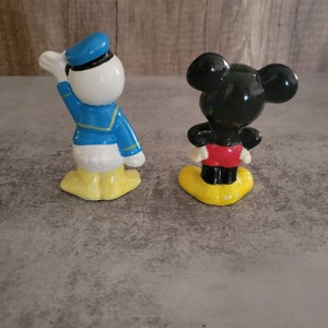 Op de afbeelding: Een vintage set keramische beeldjes met Mickey Mouse en Donald Duck. Mickey draagt een rode broek en gele schoenen, terwijl Donald een blauw matrozenpak en gele schoenen draagt.