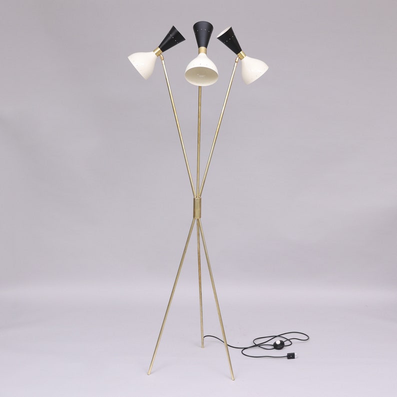 Pu&ograve; includere: Una lampada da terra a treppiede dorata con tre paralumi a forma di cono bianco e nero. La lampada ha un cavo nero e una spina nera.