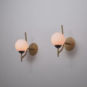 May include: Two wall sconces with a brass finish. Each sconce features a round, frosted glass globe light, a black base, and a curved brass arm attached to a circular brass wall plate. The lights are mounted on a light gray wall.