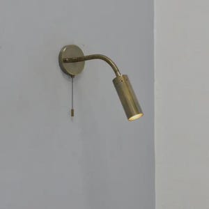 May include: A brass-toned wall-mounted reading lamp. The lamp features a circular base, a flexible gooseneck arm, and a cylindrical lamp head. A pull chain hangs from the base. The lamp is designed to be mounted on a wall for focused lighting.