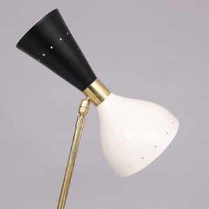 Pu&ograve; includere: Una lampada da terra regolabile in bianco e nero con un braccio in ottone e un paralume a forma di cono. Il paralume ha un motivo di piccoli fori.