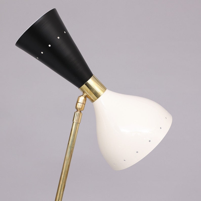 Pu&ograve; includere: Una lampada da terra regolabile in bianco e nero con un braccio in ottone e un paralume a forma di cono. Il paralume ha un motivo di piccoli fori.