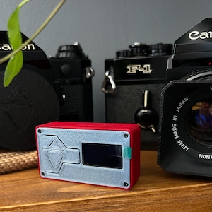 Puede incluir: Un dispositivo rojo y plateado con un logotipo de diamante y una pequeña pantalla. El dispositivo está sentado en un estante de madera junto a dos cámaras Canon negras vintage.