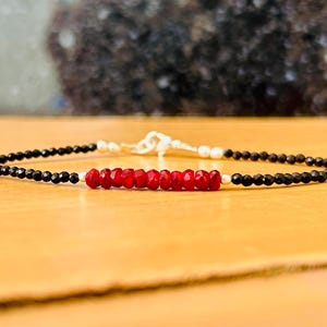 Può includere: Un braccialetto con perline nere e rosse con una chiusura argentata. Il braccialetto è realizzato con piccole perline nere e una fila di perline rosse al centro.