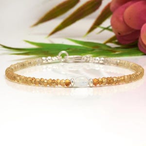 May include: A delicate bracelet with small, faceted golden-yellow beads and a central oval, milky-white gemstone. The bracelet has a silver clasp. The background is soft and blurred, with floral accents. A piece of jewellery.