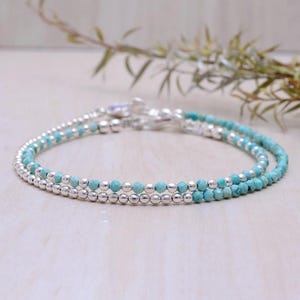 May include: Two delicate bracelets, one with turquoise beads and the other with silver beads, are layered together. The bracelets are on a light wood surface.