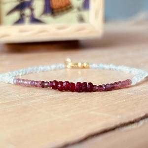 May include: A delicate bracelet with a gradient of light blue, pink, and red beads. The bracelet features a row of red gemstone beads in the center.